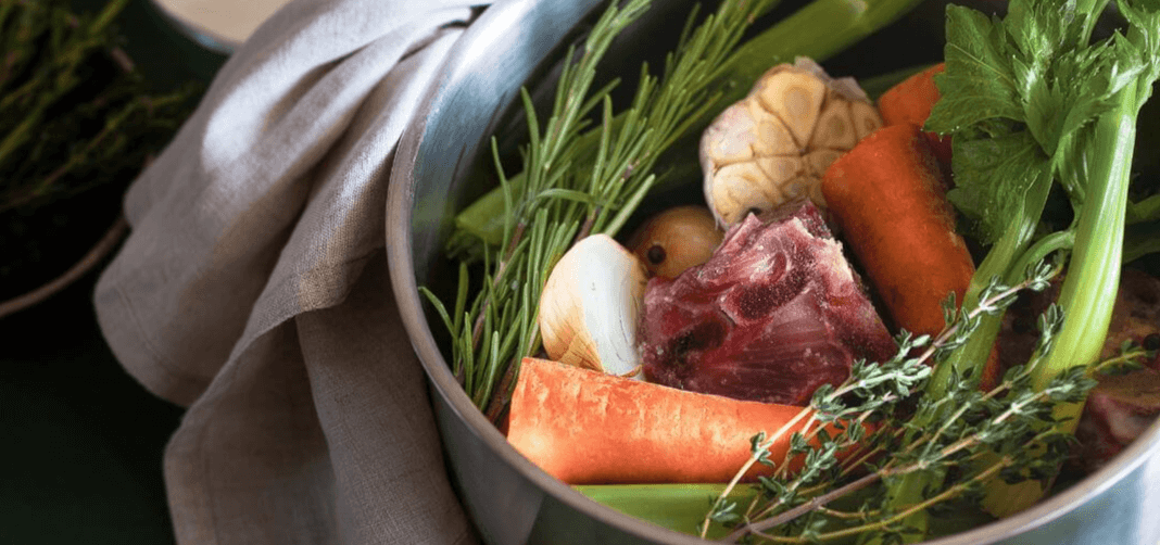  Close-up of a pot containing beef bones, carrots, celery, onions, and herbs, cooking to make bone broth.