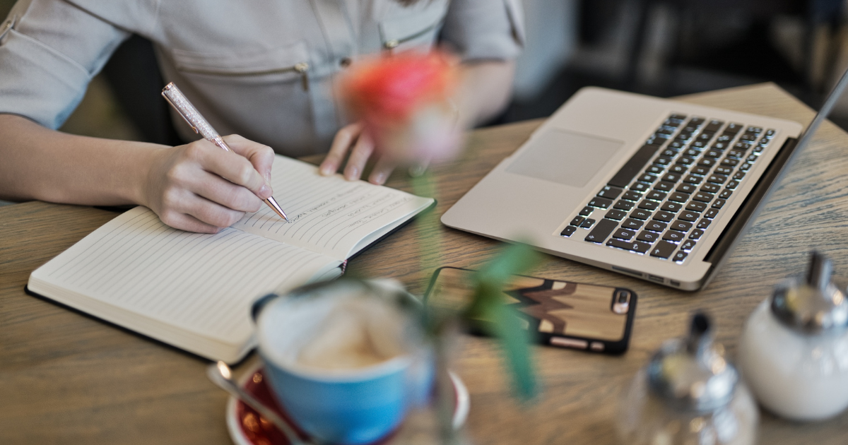 writing in journal at desk next to phone and laptop coffee and flower