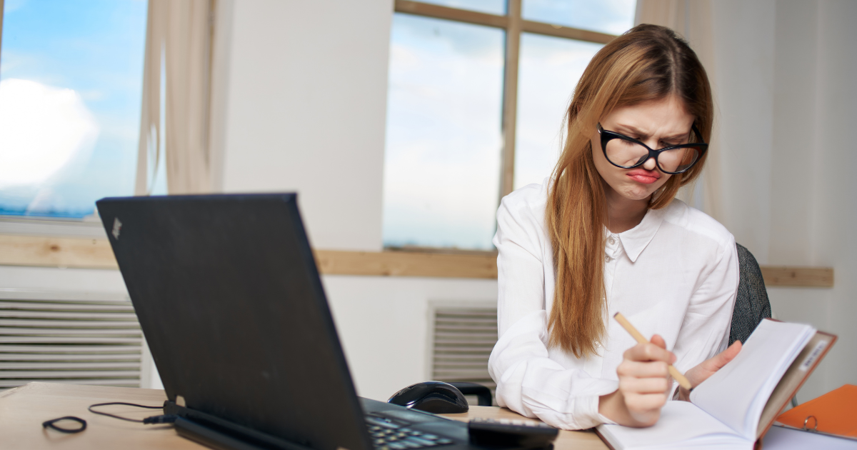 woman with glasses frustrated expression writing in journal at desk near laptop.