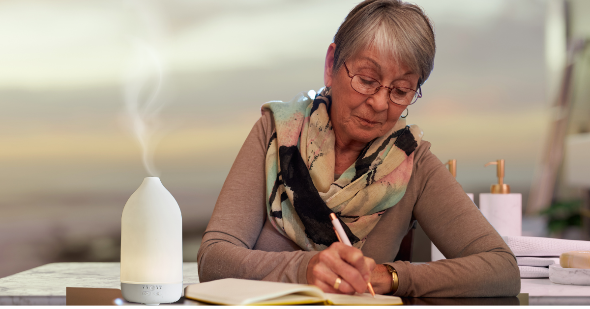 Older woman wearing glasses writing in journal at a desk with diffuser by her elbow