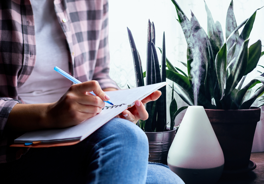 person in plaid shirt writing in journal on their knee with diffuser and plants behind