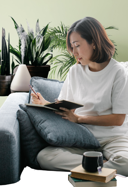 Woman sitting on couch writing in journal with diffuser and plants in background, journals and cup of tea in foreground