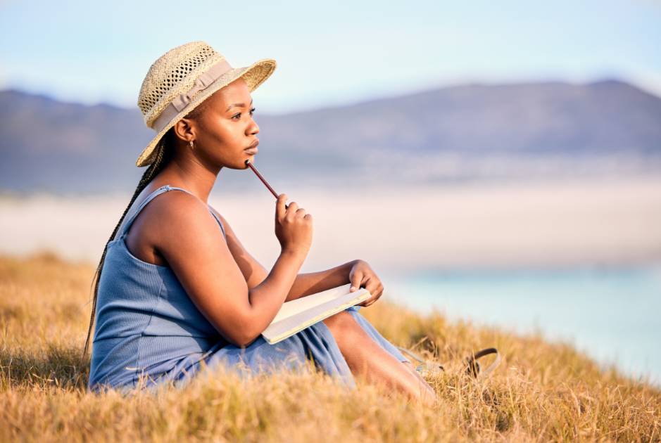 woman wearing hat sitting in the grass by a lake journaling with pencil resting on her chin