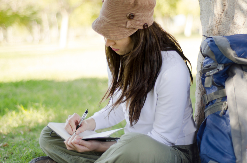 woman sitting in the grass in front of a tree journaling with a backpack behind her