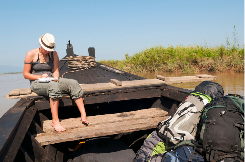 woman itting on a wooden boat in the sun journaling with backpacks and gear in the boat