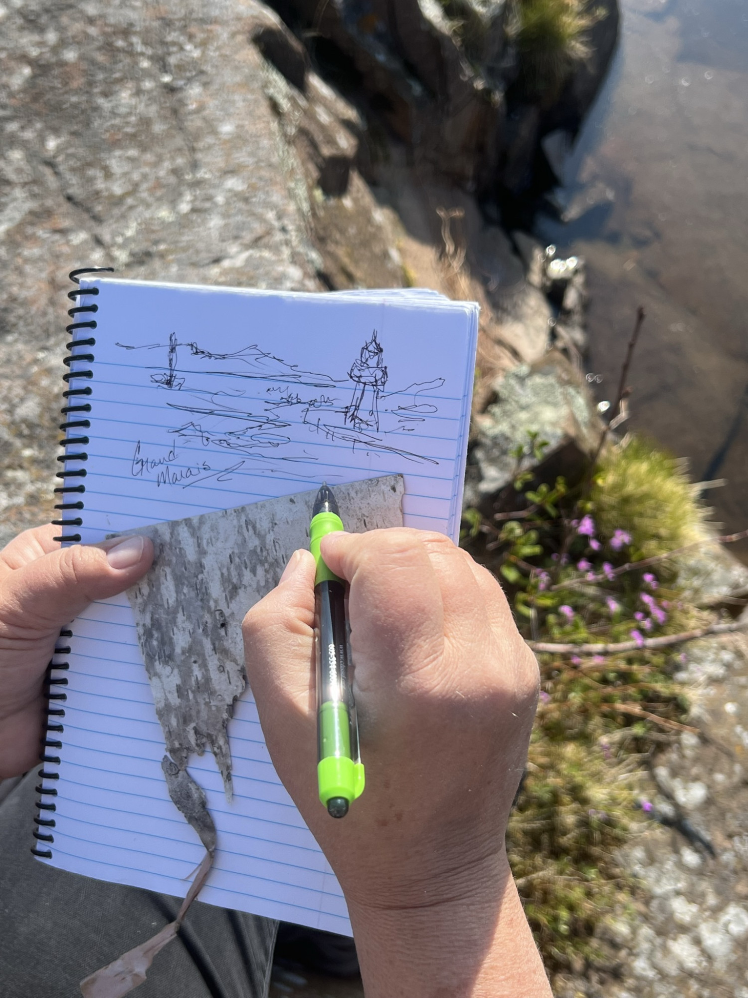 Drawing the Grand Marais lighthouse while sitting on a rock on the bank of Lake Superior.  A piece of found bark for later journal entries.