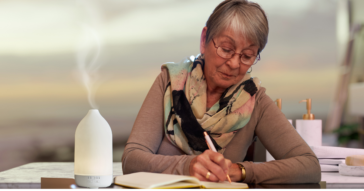 Older woman sitting at a desk journaling with diffuser on desk