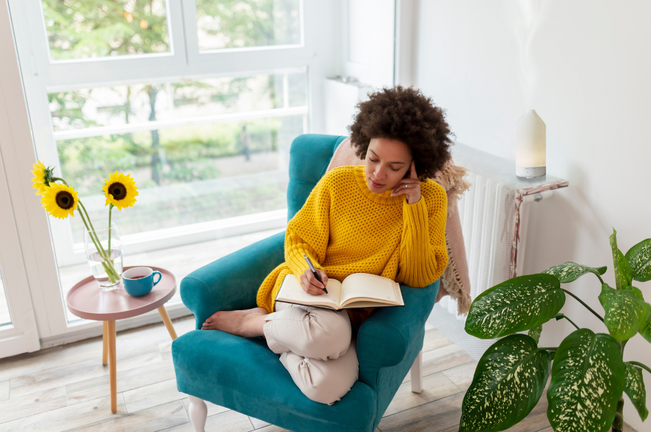 Woman sitting on a bright turquoise blue chair with sunflowers writing in her journal with diffuser behind her for blog post "Essential Aromas: Diffuser Recipes to Elevate Your Journaling Experience"