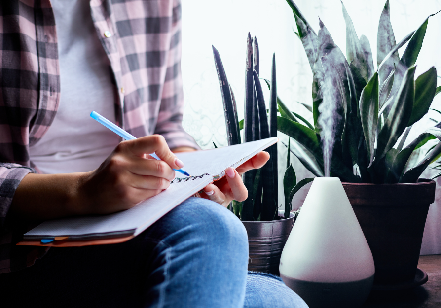 Person holding a journal on their lap, writing, a diffuser and plants in the background  for blog post "Essential Aromas: Diffuser Recipes to Elevate Your Journaling Experience"