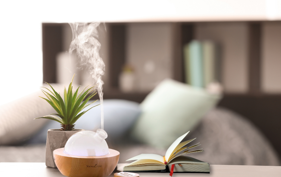 Aria diffuser next to an open journal, plant, and a bed in the background covered in pillows for blog post "Essential Aromas: Diffuser Recipes to Elevate Your Journaling Experience"