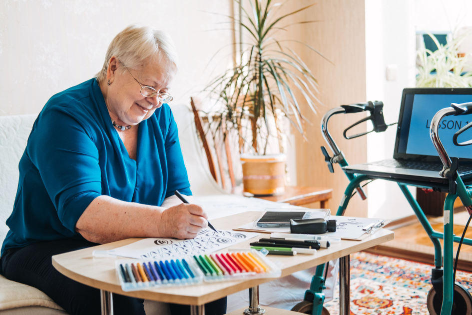 Older woman at a table working on an image in her journal using markers, her walker sits in the backgroud with a plant and laptop screen image for post Why Take an Introductory Class for Journaling? 