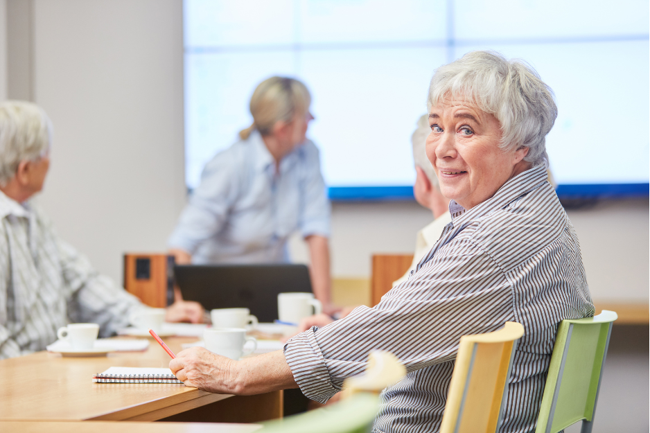 older woman in a group room setting at a desk looking over her shoulder and smiling, coffee cups and journals on the table, image for post Why Take an Introductory Class for Journaling? 