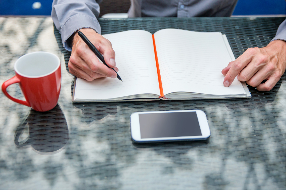 older male hands about to write in journal, bright red-orange coffee cup, and cell phone on outdoor table for post Impact vs goals