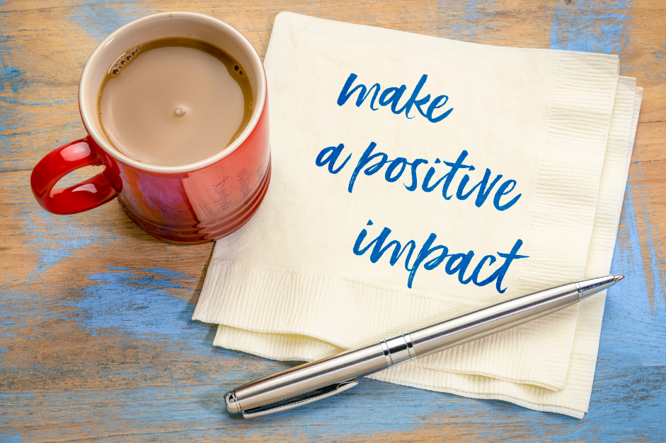 A bright red-orange coffee cup, pen, and table napkin on blue worn table. the words 'Make a positive impact' is written on the napkin for post about impact vs goal