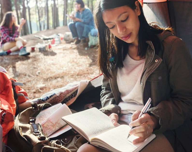 Woman sits in open door of tent writing in her journal. background is forest and two other people having beverages and sitting on the ground image for impact vs goals
