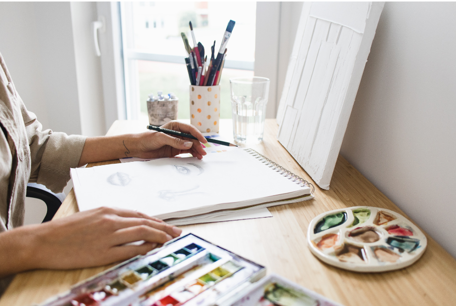Person sitting in front of a journal sketch book holding a pencil next to a bright window, water colors, and palette for post Creatively Unlock Your Thoughts with Image-Based Journaling!