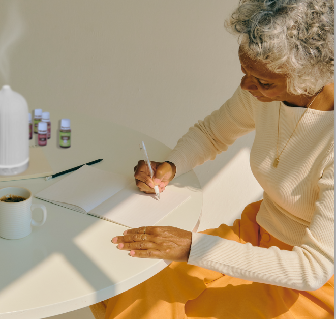Woman with graying hair and bright mustard colored pants is sitting at a table in the sunlight writing in a journal with a cup of a beverage, diffuser, and essential oils on the table image for post Journaling as a form of meditation: How essentail oils support the experience