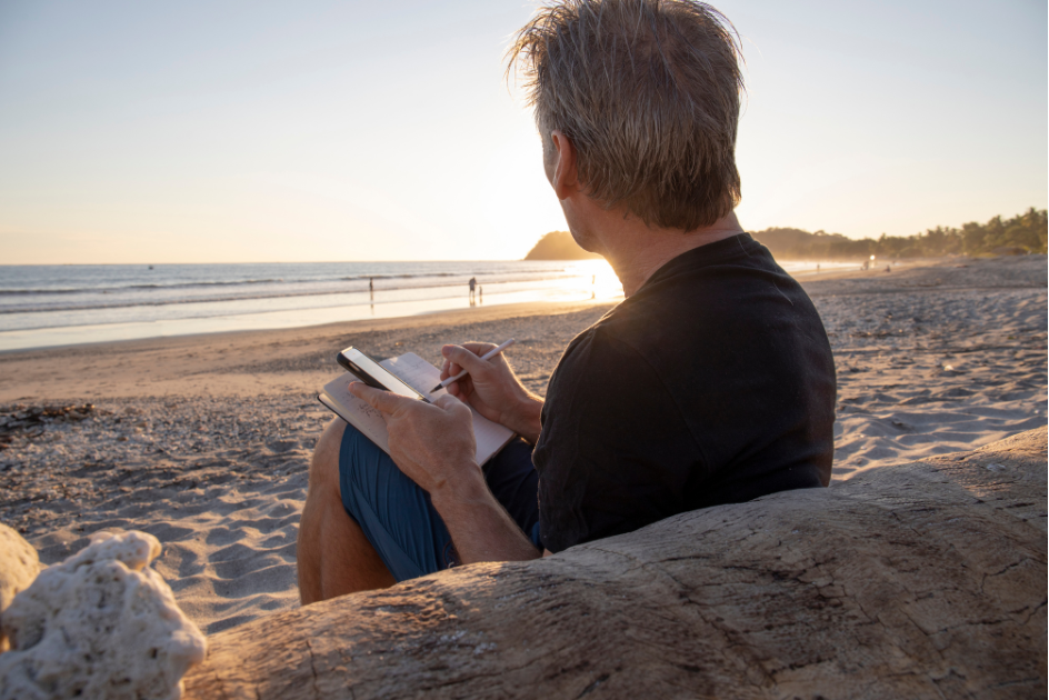 Person sitting on the beach looking toward the water with a journal in their hand image for post Journaling as a form of meditation: How essentail oils support the experience