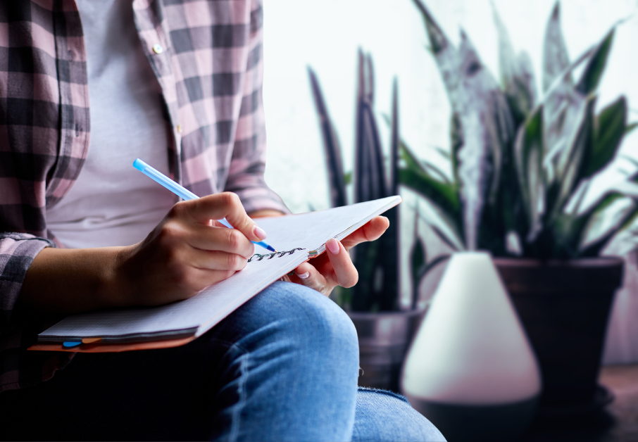 Person sitting with knees crossed writing in a journal with plants and a diffuser behind them image for post Journaling as a form of meditation: How essentail oils support the experience