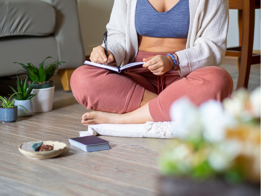 Woman sitting on a pillow on the floor in a living room with plants near her, her journal is on her knee and she's writing image for post Journaling as a form of meditation: How essentail oils support the experience