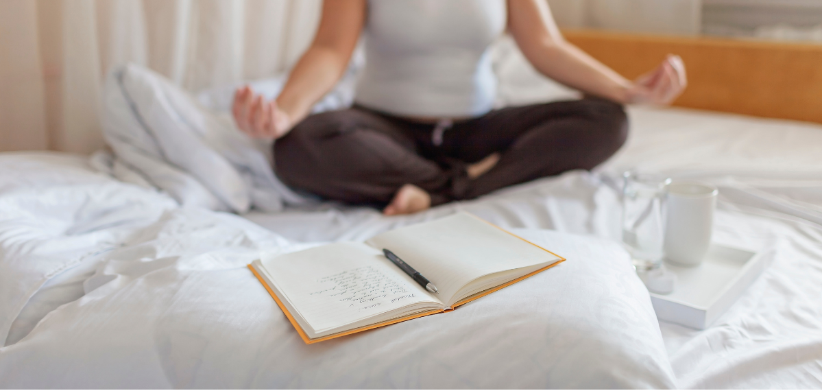 woman sitting on a bed in a meditation pose with her journal and pen on the bed for a post titled Journal Prompts to Unlock Creativity, Clarity, and Focus!