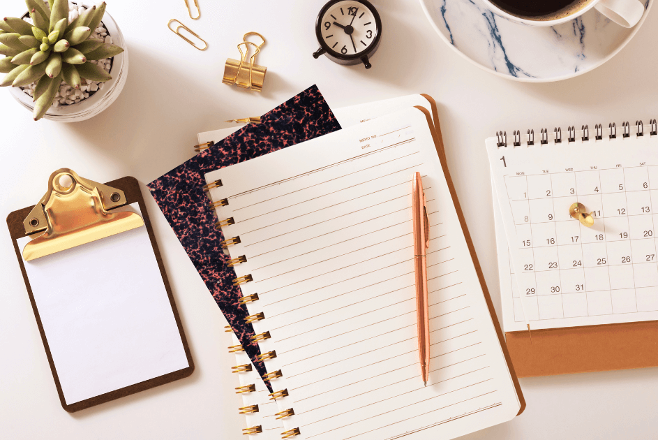 Three stacked notebook style journals with a pen on top.  A clipboard, open calendar, plant, cup of coffee and paper clips sit next to it on the table for a post titled4 Types of Journals: Which One Fits Your Style? 
