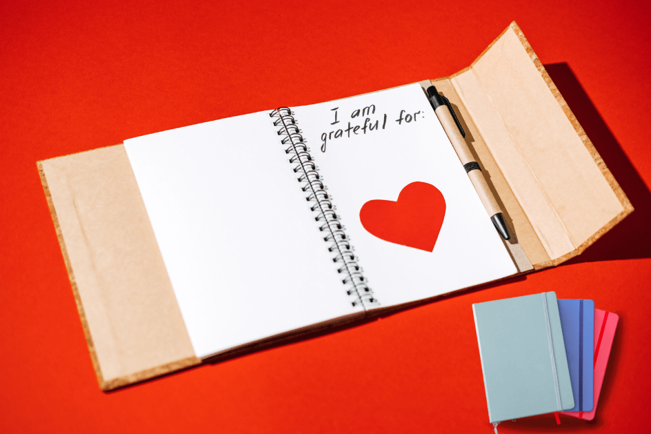 An open gratitude journal. Journal is a tri-fold design and page has a large red heart, plus there are 3 mini journals in front of that for for a post titled4 Types of Journals: Which One Fits Your Style? 