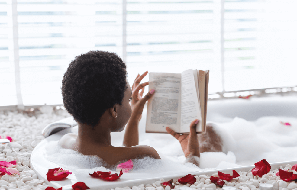 Woman sitting in a bubble bath reading a book next to a window and rose petals everywhere for a post titled How to Prioritize Self-Care in a Hectic Day