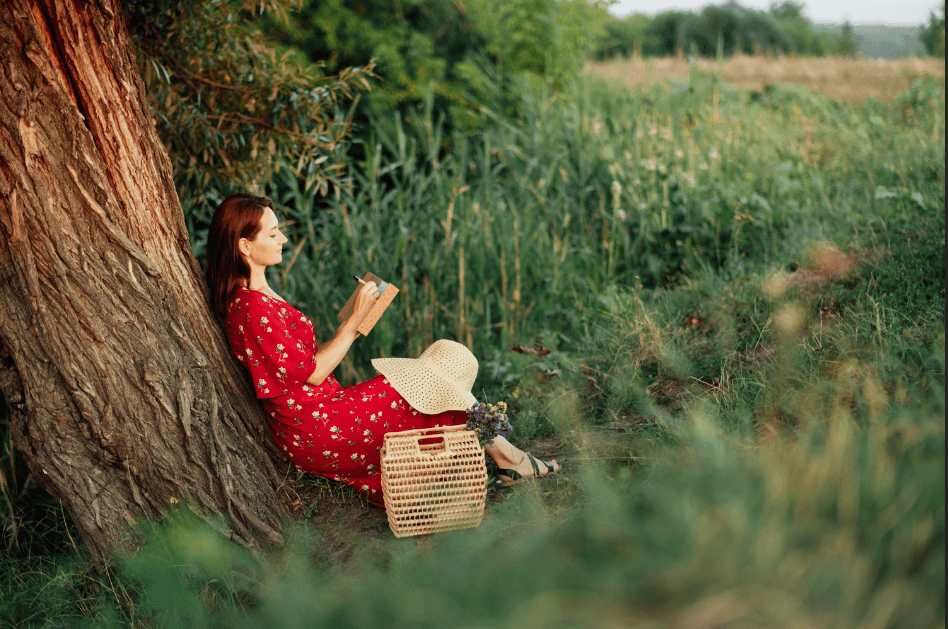 Woman wearing a red dress with her bag and had by her leg, leaning against a tree, writing in her journal for blog post titled How to Prioritize Self-Care in a Hectic Day