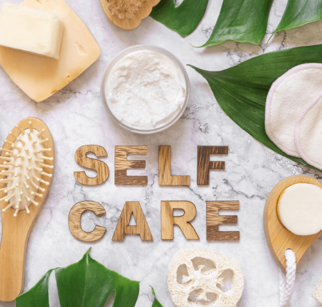 Wooden letters spelling SELF CARE are laying on a marble top table next to luffa sponge, pumas stone, hair brush, leaves, and other spa items for post titled How to Prioritize Self-Care in a Hectic Day