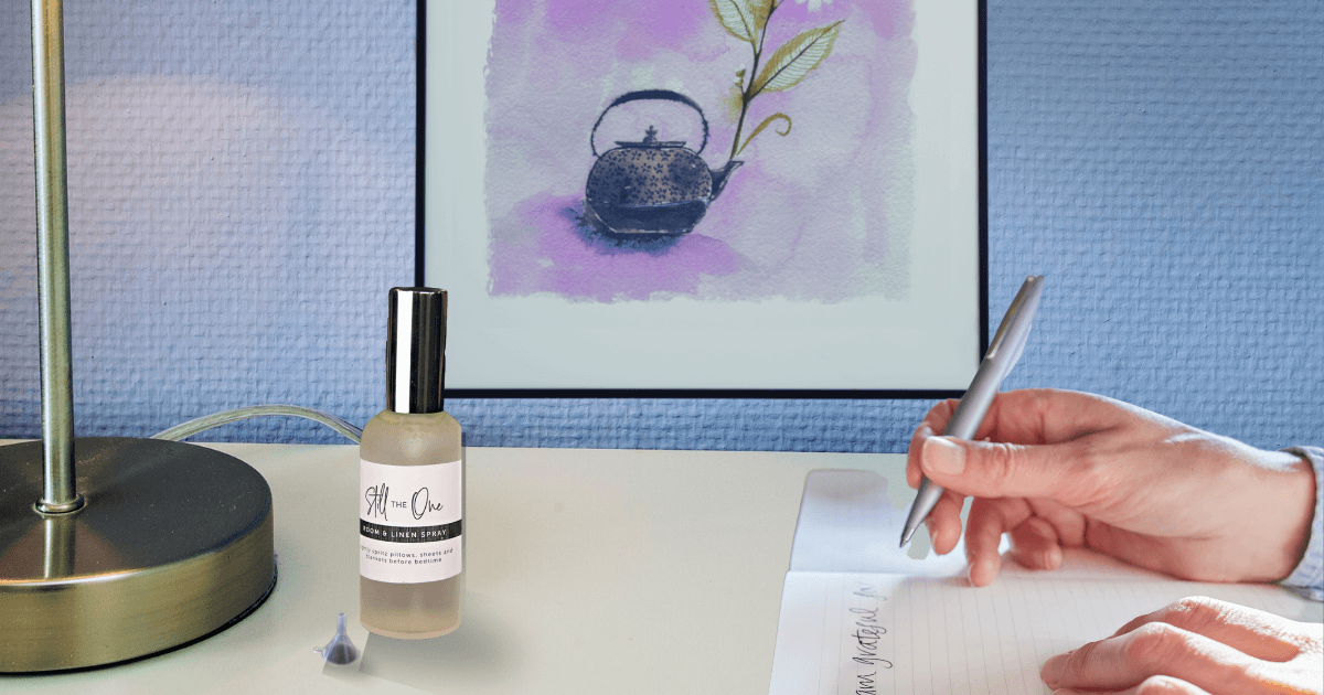 A bottle of Linen spray on a night stand next to a lamp, painting in the background on a blue wall, hands coming in from the right writing in a journal A bottle of Myrrh, Peace & Calming, and Clary Sage essential oils for blog post titled Linen Spray DIY: "You're still the one!"