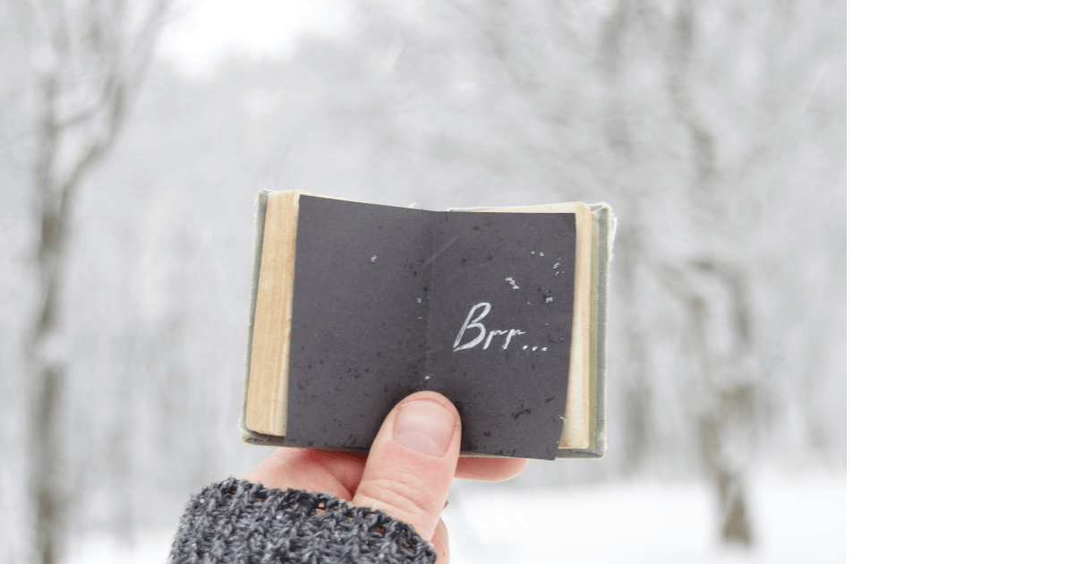 snow background, hand holding a mini journal open to black page with Brr... written in chalkfor blog post titled 10 Journal Prompts to Help Harness Your Energy 