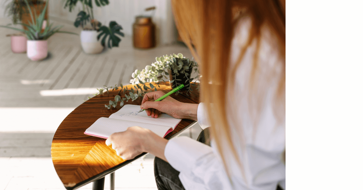 Person sitting at a small desk writing in a journal with light streaming in from a window and lots of plants in the are  for a post titled How House Plants Can Enhance Your Journaling Experience 