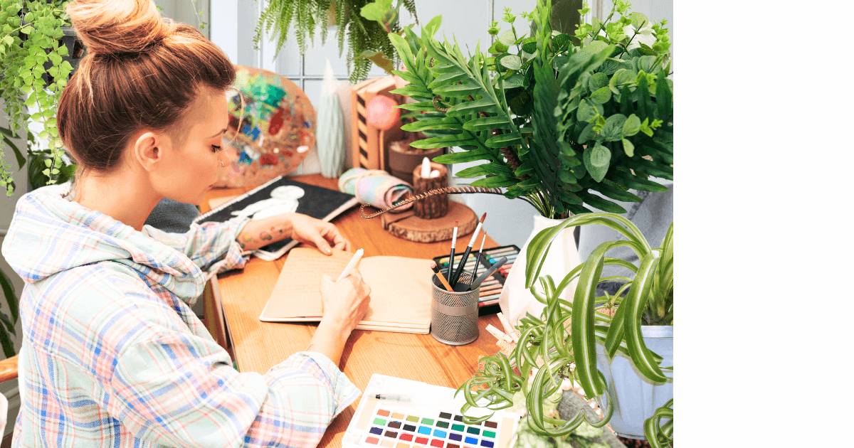 woman sitting at a wood desk journaling with art supplies and plants all around her for blog post titled How House Plants Can Enhance Your Journaling Experience