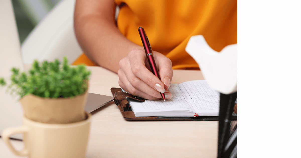 Person in saphron colored shirt writing in a journal at a desk with a small plant in a coffee cup for post titled  How House Plants Can Enhance Your Journaling Experience