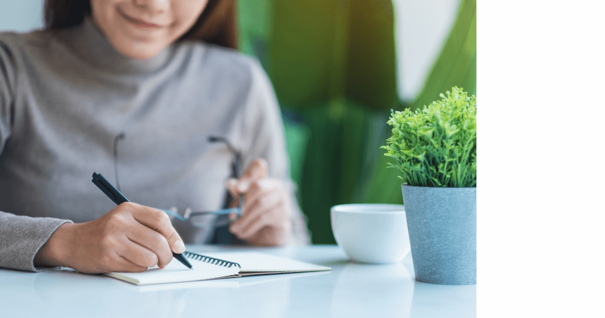 person sitting at a desk writing in a journal next to a cup and plants for post titled How House Plants can enhance your journaling experience