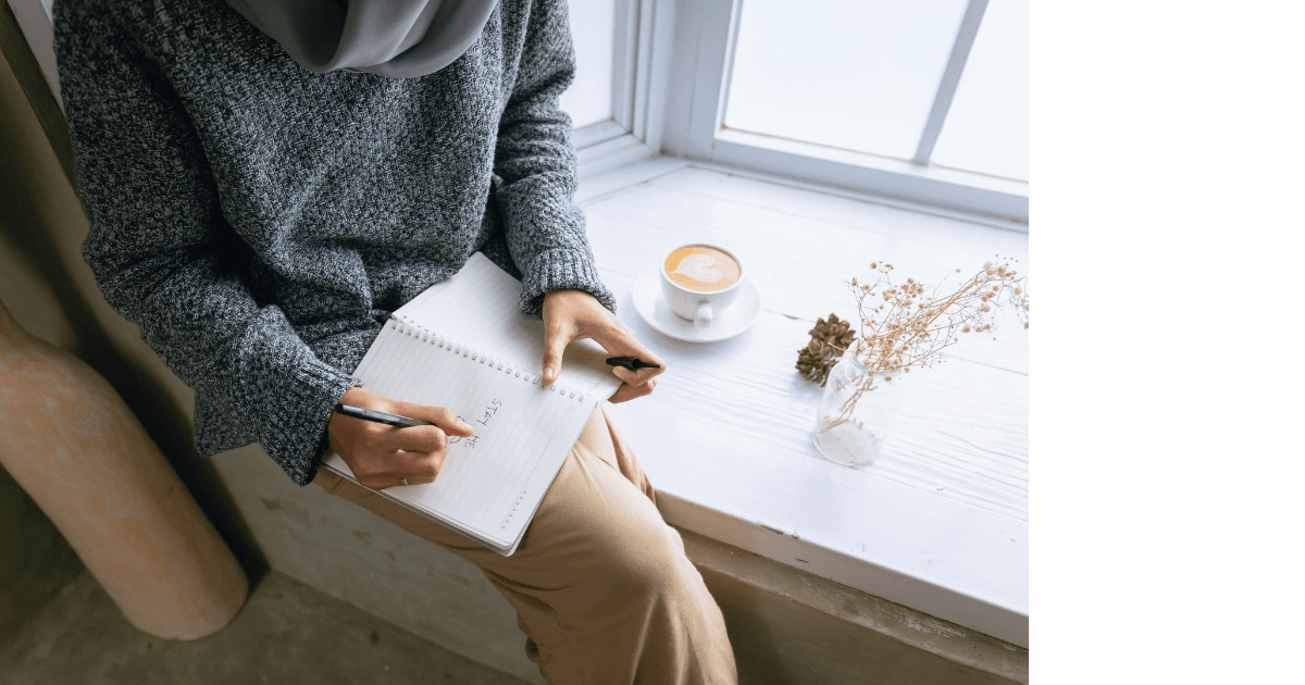 person wearing a big sweater sitting in a window seat and writing in a journal, coffee cup on the ledge, dried flowers on ledge near window for post titled 5 Tips to Get Your Mind Off Winter and Thinking Spring!