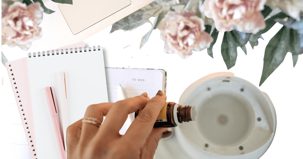 A hand is dropping Young Living essential oils into an open diffuser, in the background are light pink journals, a pink pen, and pink peonies for post titled Spring Aromatherapy: Refreshing Diffuser Recipes