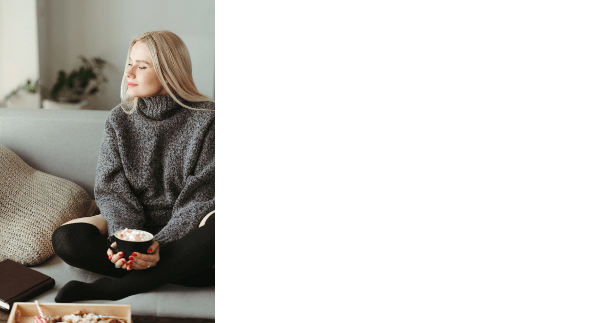 woman sitting crosslegged on couch with closed eyes facing the sun coming in the window while holding a warm cup of something for post titled Embrace Hygge with Journaling and Essential Oils