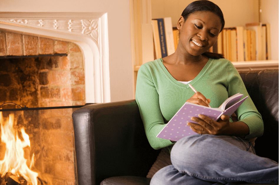 Woman sitting on a couch next to a fireplace journaling with a smile on her face for post titled Embrace Hygge with Journaling and Essential Oils