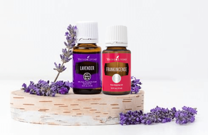 a bottle of lavender essential oil and a bottle of frankincense essential oil sitting on a slice of birch bark with fresh lavender laying around for a post titled DIY "Ouch Relief" Roller Ball Recipe