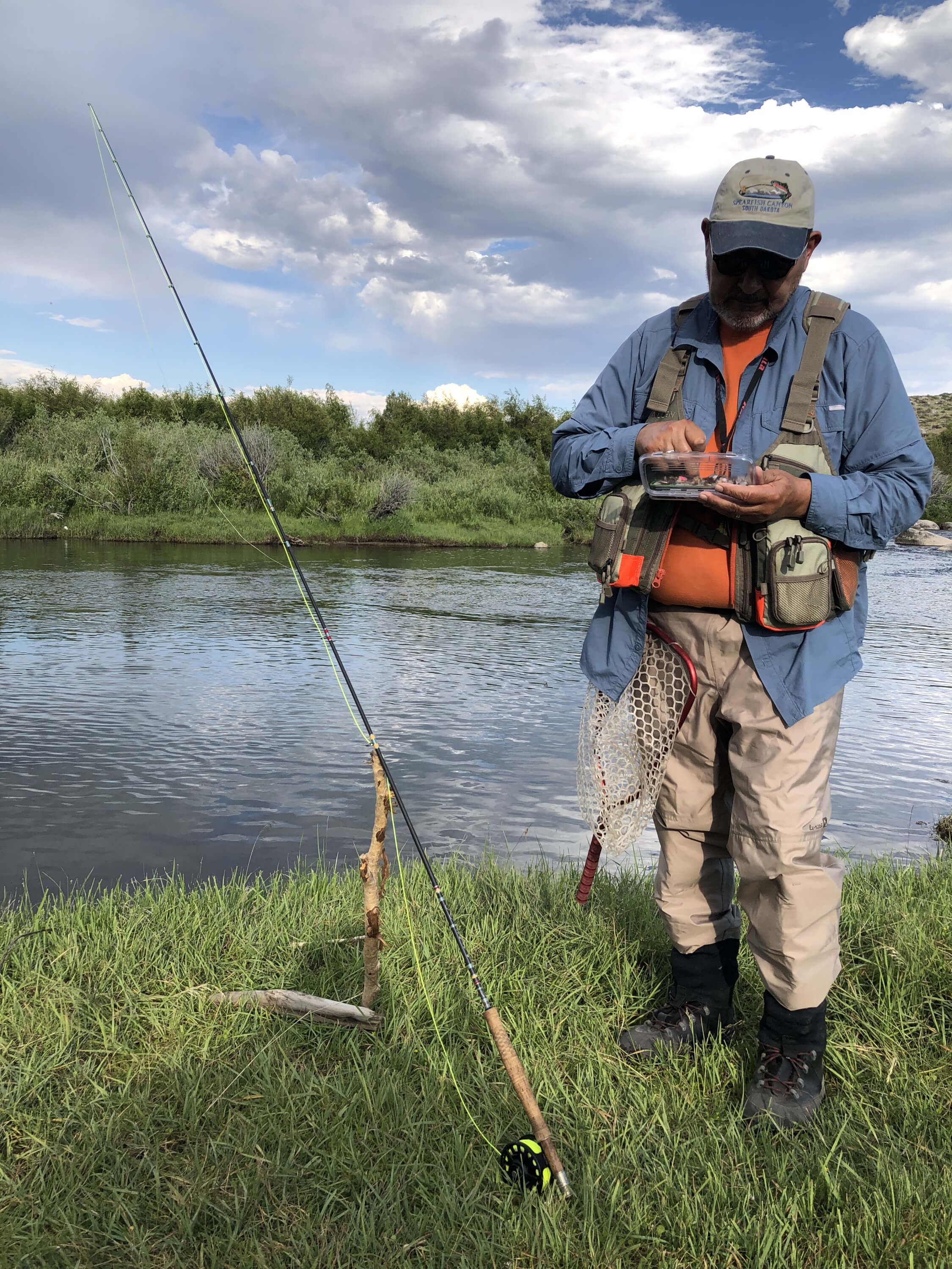 Allen standing on the lake shore wearing his fishing waders, his pole on the ground, as he rummages through his fly fishing lures for a blog post titled Reeling in the Benefits of a Fishing Journal