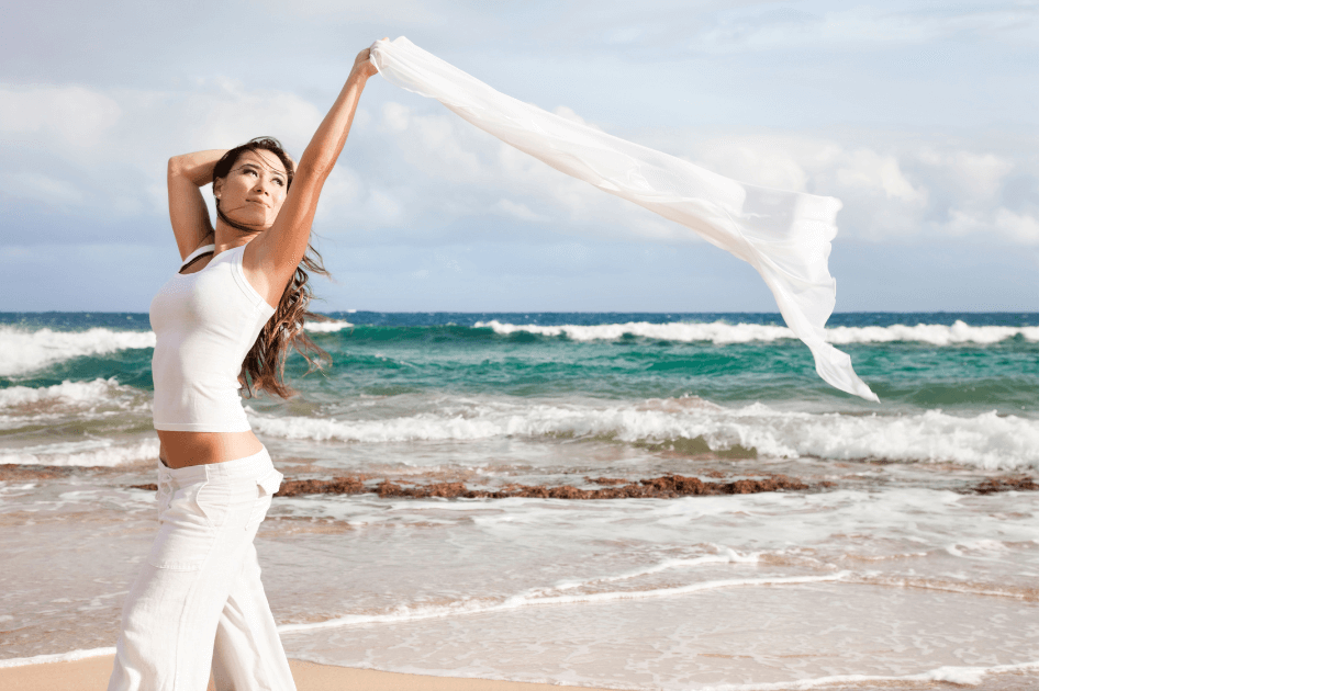 Woman wearing white holding a white scarf over her head that's blowing in the wind as she walks the sandy shore of an ocean with waves behind her for a blog post titled Summer Aromatherapy: 5 Diffuser Recipes