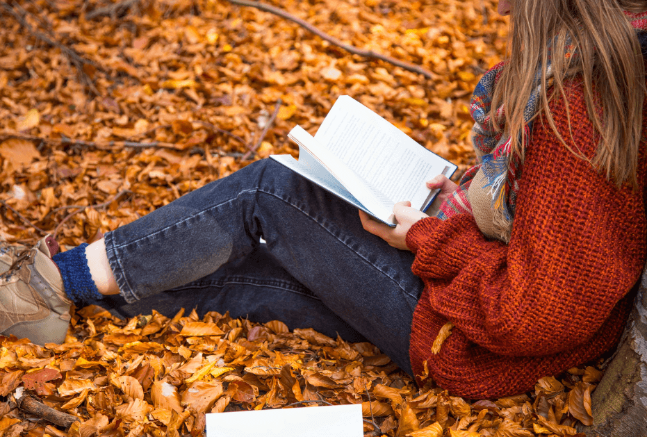 Person wearing a rust colored big sweater, jeans, and boots sitting on the ground thats covered in fall leaves leaning up against a tree and writing in her journal or a Bound to Journal blog post titled Fall Aromatherapy: 5 Seasonal Diffuser Recipes 