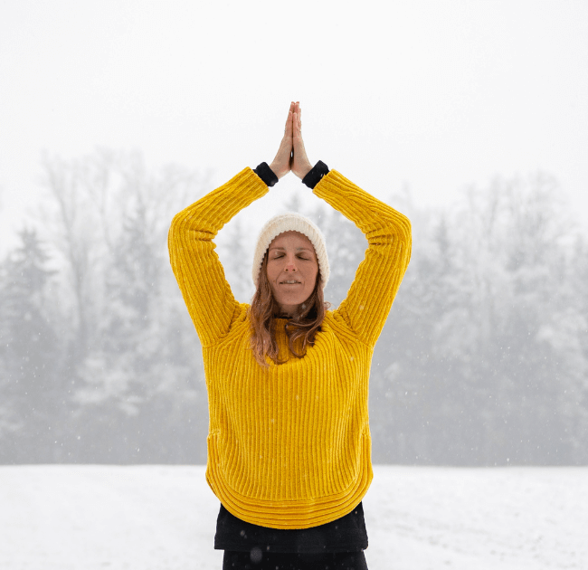 Person outside in winter wearing a bright mustard colored sweater with her arms above her head in a yoga pose  for a blog post titled Winter Diffuser Recipes for Clarity, Inspiration, and Cozy Productivity