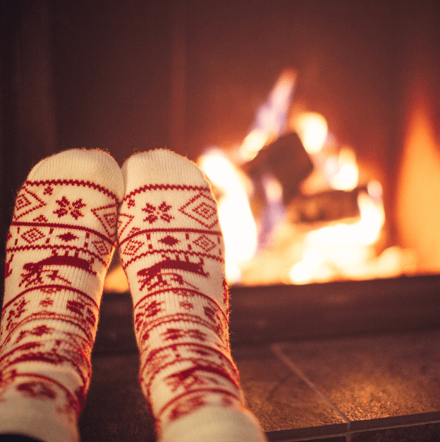 A pair of feet wearing Scandinavian styled red and white winter socks with a roaring fire showing behind them  for a blog post titled Winter Diffuser Recipes for Clarity, Inspiration, and Cozy Productivity