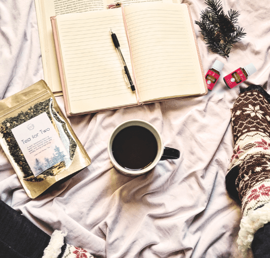 There's an open journal, coffee cup, bag of tea, and essential oils sitting on a blanket with just a bit of two legs showing that are wearing fuzzy patterned winter socks  for a blog post titled Winter Diffuser Recipes for Clarity, Inspiration, and Cozy Productivity