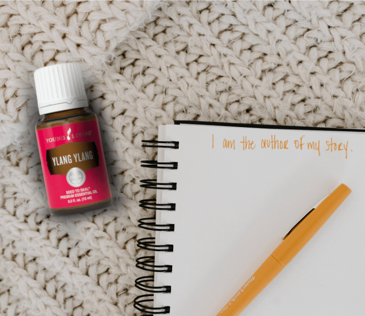 A bottle of Young Living's Ylang Ylang is laying on a white sweater type throw next to an open journal and pencil   for a blog post titled Winter Diffuser Recipes for Clarity, Inspiration, and Cozy Productivity