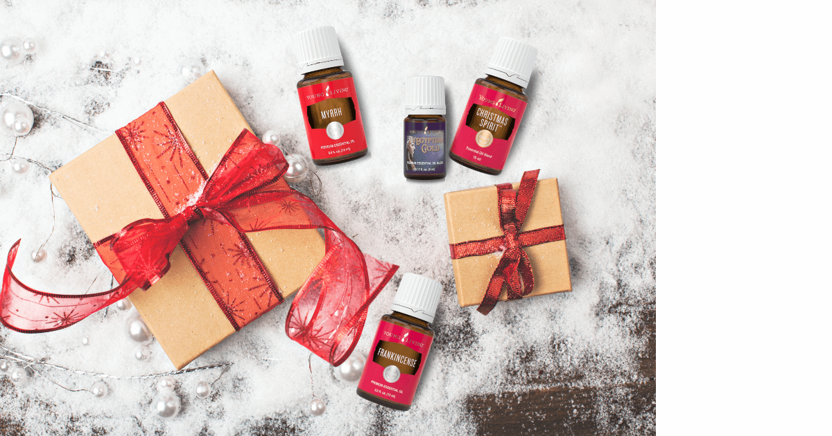 Four essential oils (Frankincense, myrrh, egyptian gold, and Christmas spirit) and two craft paper wrapped gifts tied in red ribbon laying on snow for a blog post titled Ancient Oils: The Perfect Companion for Holiday Journaling