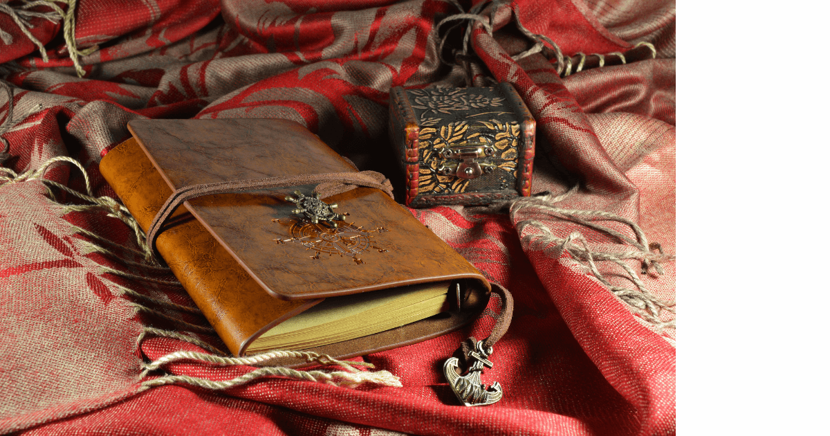  An old leather bound journal with bookmark hanging out laying on a gold and orange-red  scarf with a little wooden carved box  A bottle of young living myrrh essential oil near a gold christmas ornament and branches from an evergreen in the background for a blog post titled Ancient Oils: The Perfect Companion for Holiday Journaling